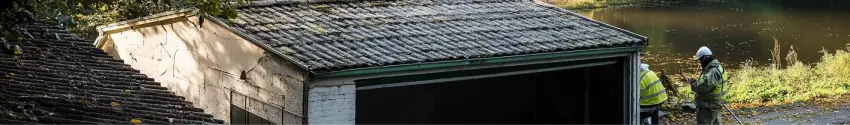 Two workmen examine a small building with an asbestos roof next to a river